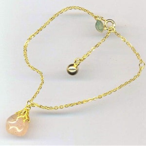 Puede incluir: Un delicado brazalete de cadena dorada con un colgante de piedra rosa en forma de l&aacute;grima y un peque&ntilde;o abalorio de bola dorada.