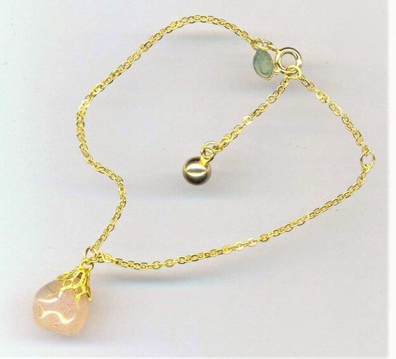 Puede incluir: Un delicado brazalete de cadena dorada con un colgante de piedra rosa en forma de l&aacute;grima y un peque&ntilde;o abalorio de bola dorada.