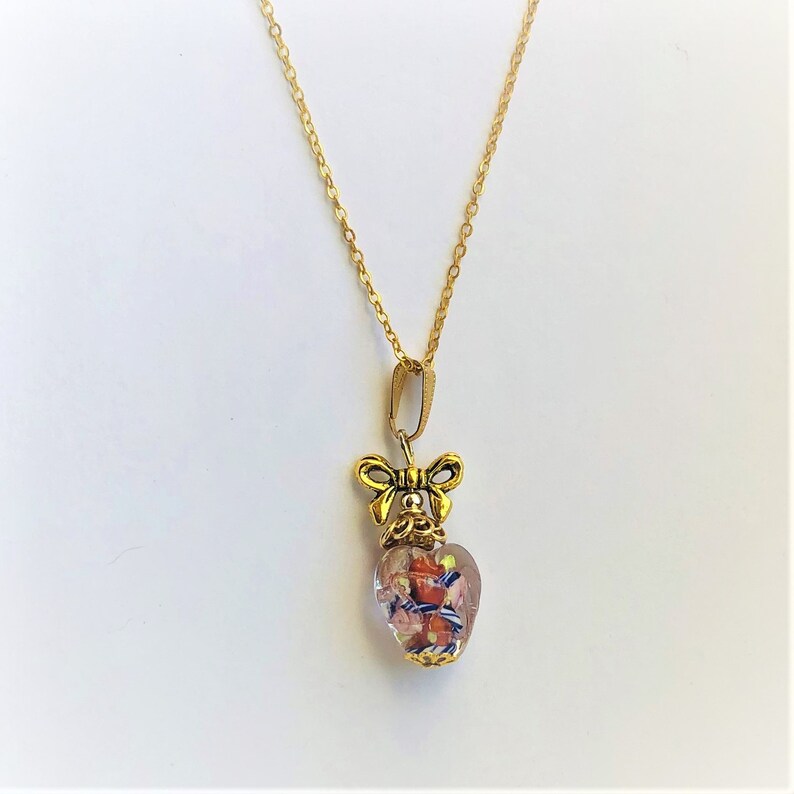 May include: A gold chain necklace with a heart-shaped pendant. The pendant is made of clear glass with colorful flecks and a gold bow on top.