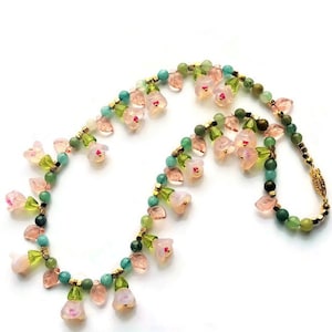 May include: A delicate necklace featuring pink glass flowers, green leaves, and gold beads. The necklace is made with a variety of natural stones, including jade and hematite.
