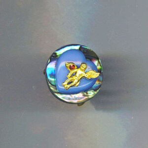 May include: A blue heart-shaped button with a gold cherub design and a red gemstone accent. The button is made of abalone shell with iridescent colors.