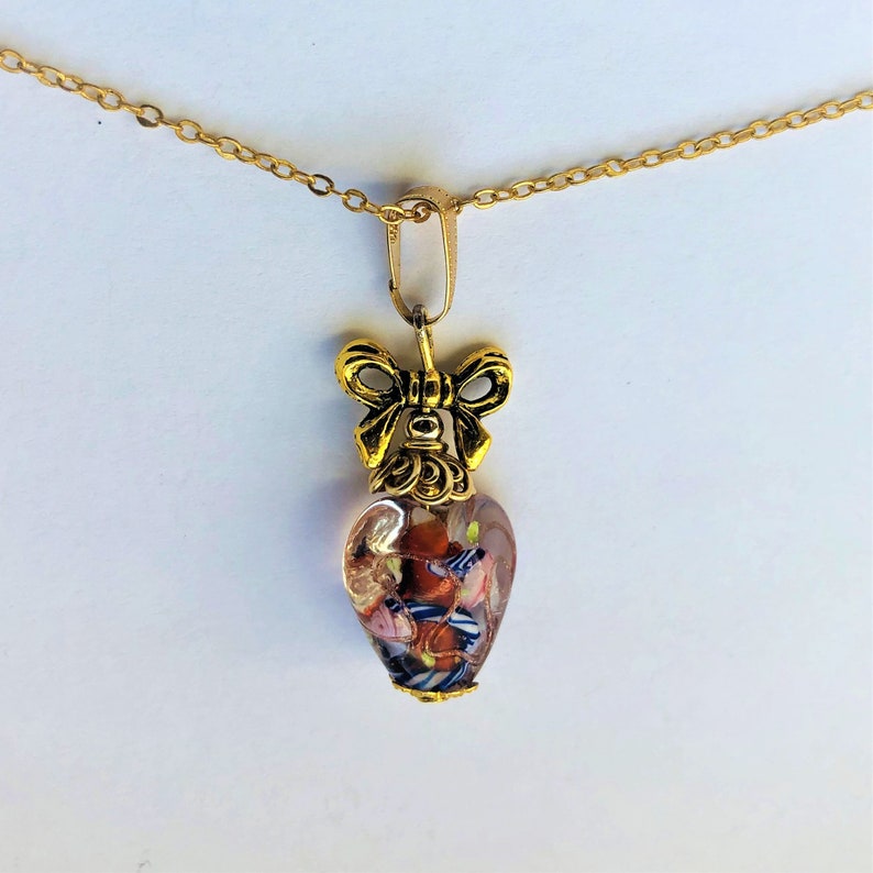 May include: A gold chain necklace with a heart-shaped pendant. The pendant is made of clear glass with colorful flecks and a gold bow at the top.
