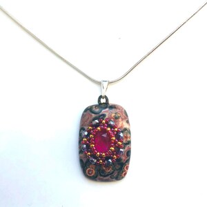 May include: A silver chain necklace with a rectangular pendant featuring a pink, brown, and black patterned stone with a red gemstone surrounded by smaller purple and gold beads.