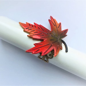 May include: A red, orange, and green metal maple leaf napkin ring. The ring is decorated with a gold-colored design.