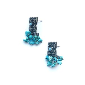 May include: A pair of silver-toned earrings with a rectangular top and dangling turquoise stones. The earrings have a textured design on the top portion and are set against a white background. The turquoise stones are various shapes and sizes.