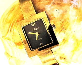 May include: A gold-toned wristwatch with a square face and a black dial. The watch face has gold-toned hands and markers, and the text "GARDEN" and "QUARTZ" is printed on the dial.