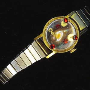 May include: A gold-tone wristwatch with a clear face and red jewels. The watch has a silver metal band.