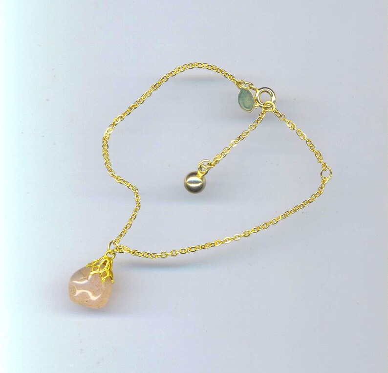 Puede incluir: Un brazalete de cadena dorada con un colgante de piedra rosa y un peque&ntilde;o acento de piedra verde. El brazalete tiene una longitud ajustable.