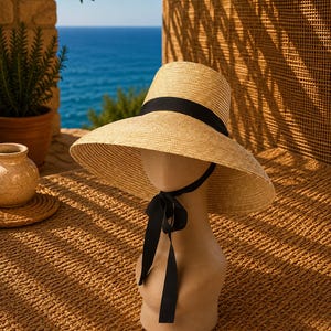 May include: A wide-brimmed straw hat featuring a natural tan woven texture and a solid black ribbon band encircling the crown. The hat includes a black ribbon chin strap that ties into a bow beneath the chin, providing a secure fit for the wearer.