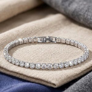 May include: A silver tennis bracelet featuring a continuous line of clear, round-cut gemstones. The bracelet is displayed on a neutral fabric. The clasp is visible, highlighting the design and how it fastens. A classic piece of jewellery.