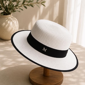May include: A white straw hat with a flat top and a wide brim featuring a black trim along the edge. A black ribbon band encircles the base of the crown, adorned with a centred silver-toned letter M emblem. The hat has a structured, woven texture.