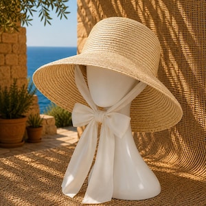 May include: A light beige woven straw sun hat with a wide brim and a smooth white fabric ribbon tied into a bow under the chin. The hat is displayed on a plain white mannequin head, showcasing the textured material and the long, flowing ribbon ends.