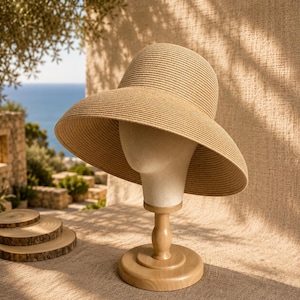 Può includere: Un cappello da sole in paglia color marrone chiaro con una tesa larga inclinata verso il basso e una corona arrotondata. Il cappello presenta una trama finemente intrecciata su tutta la superficie, con una forma strutturata su un supporto.