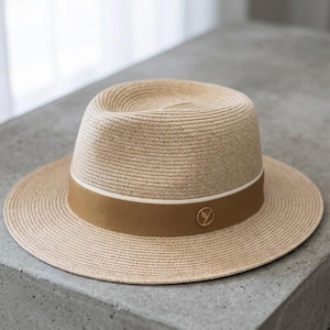 Pode incluir: Um chapéu fedora bege com uma faixa marrom e um emblema dourado. O chapéu tem uma aba larga e uma coroa estruturada. A faixa é acentuada com uma fina faixa branca. O chapéu é feito de material tecido.