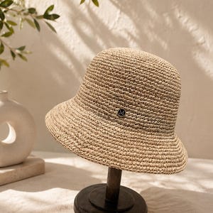 Puede incluir: Un sombrero de pescador de paja trenzada en color beige con una copa redondeada y un ala inclinada hacia abajo. Un pequeño emblema circular de metal negro con la letra M en blanco está centrado en la parte frontal, justo sobre el ala.