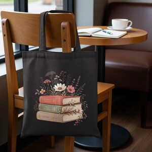 May include: A black tote bag featuring a graphic print of three stacked books in shades of red, green, and beige. The books are adorned with illustrations of various wildflowers and foliage, with the words Botanical Sketches, Wild Flowers, and Poems.