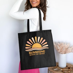 Può includere: Borsa tote nera con una grafica solare retrò e la scritta "SUNSHINE STATE OF MIND". Il design utilizza una calda tavolozza di colori. La borsa ha lunghi manici neri.