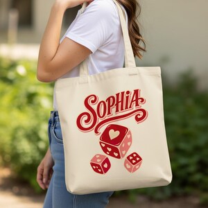 May include: A cream-colored canvas tote bag featuring the name Sophia in large red script lettering above three red dice decorated with small heart patterns. The bag has two shoulder straps and is made of a durable, lightweight fabric material.
