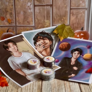 Peut inclure: Trois impressions d'art rectangulaires présentant des illustrations numériques de personnages, avec deux rouleaux de ruban adhésif décoratif placés au centre. Les impressions montrent des portraits détaillés, dont un avec le texte "ARMY" sur les doigts.