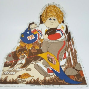 May include: A fabric applique pattern featuring a boy holding a football, a dog, and a flag with the text "Cabbage Patch Kids". The pattern is on a white background with brown, blue, red, yellow, and green accents. The pattern is designed to be sewn into a stuffed toy.