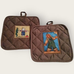 May include: Two brown quilted pot holders with a quilted diamond pattern. One pot holder features a scarecrow with a pumpkin and the other features a pumpkin-headed scarecrow.