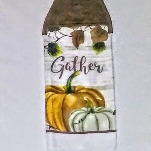 May include: A white kitchen towel with a brown hanging loop. The towel features a white background with a woodgrain pattern and the word "Gather" in brown script. A large orange pumpkin and a smaller white pumpkin are in the bottom half of the towel.