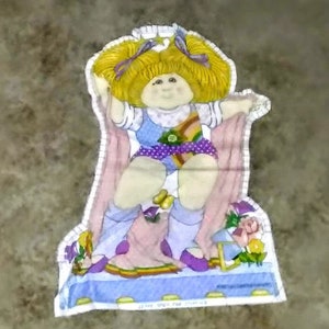 May include: A colorful fabric applique of a Cabbage Patch Kid doll wearing a purple and yellow dress with a pink cape. The doll is sitting on a blue and yellow platform with flowers and a star.