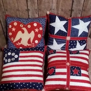 May include: Four decorative throw pillows with patriotic themes. The pillows are red, white, and blue with stars, hearts, and an eagle design. The pillows are made of fabric and have a quilted appearance.