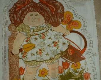 Op de afbeelding: Een stoffen patroon voor een Cabbage Patch Kid pop. De pop draagt een groen-witte bloemenjurk en houdt een gele gieter vast. De pop staat in een tuin met bloemen en een koolveld.