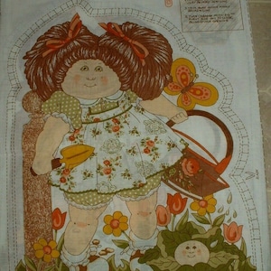 May include: A fabric pattern for a Cabbage Patch Kid doll. The doll is dressed in a green and white floral dress and holding a yellow watering can. The doll is standing in a garden with flowers and a cabbage patch.