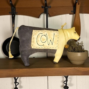 May include: A plush cow figurine with a gray body and yellow head. The word "COW" is embroidered on a rectangular patch. A small bell hangs from a gold-colored string around its neck. The cow is displayed on a wooden shelf.