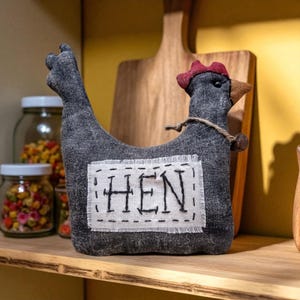 May include: A gray fabric hen-shaped doorstop with a red comb and brown beak. The word "HEN" is stitched on a white patch on the hen's body. A wooden cutting board is in the background.