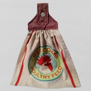 May include: A hanging kitchen towel with a red and white gingham top and a cream-colored body. The body features a circular design with a rooster graphic and the words "RED COMB POULTRY FEED" in a vintage style. Red stripes run down the sides.