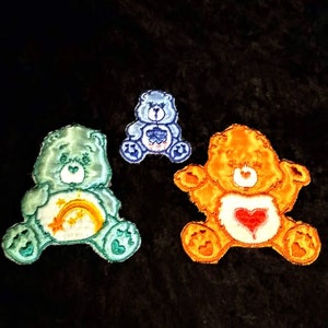 May include: Three Care Bear character iron-on patches. The patches are blue, green, and orange. The blue Care Bear has a blue heart on its chest. The green Care Bear has a rainbow and a star on its chest. The orange Care Bear has a red heart on its chest.
