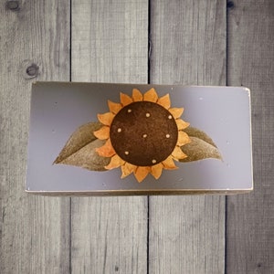 May include: A wooden sign with a painted sunflower on a gray background. The sunflower has yellow petals and a brown center with white dots.