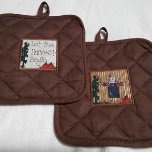 May include: Two brown quilted pot holders with a quilted diamond pattern. One pot holder has a white patch with the text "Let the Harvest Begin" and a red and orange pumpkin. The other pot holder has a patch with a cartoon scarecrow in a brown field.