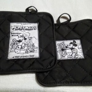 May include: Two black quilted pot holders with white Mickey Mouse designs. One pot holder features Mickey Mouse as a steamboat captain with the text "Mickey Mouse Presents Steamboat Willie A Walt Disney Comic". The other pot holder features Mickey Mouse steering a ship's wheel.