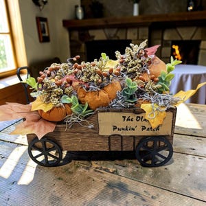 May include: A decorative wooden wagon with black wheels, filled with orange fabric pumpkins, artificial green eucalyptus leaves, brown berry clusters, and dried autumn leaves. A sign on the side reads The Olan Punkin' Patch in black lettering.