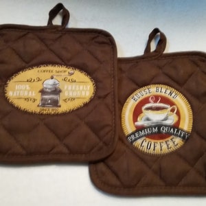 May include: Two brown quilted pot holders with a coffee theme. One pot holder features a coffee grinder with the text "Coffee Shop 100% Natural Freshly Ground Since 1919". The other pot holder features a cup of coffee with the text "House Blend Premium Quality Coffee".