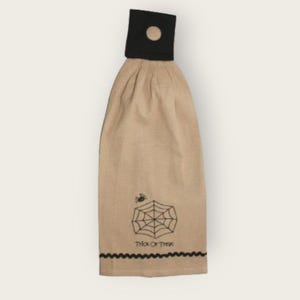 May include: A tan kitchen towel with a black spider web and spider design. The towel has a black hanging loop with a button and says "Trick or Treat" at the bottom.