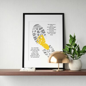 Op de afbeelding: Ingelijste kunstafdruk met een grijs en geel voetstappenontwerp en motiverende tekst. Het kunstwerk bevat een schoenafdruk en een gestileerde hand, met inspirerende zinnen in een modern lettertype. De print wordt tentoongesteld op een plank met een gouden lamp en een plant in pot.