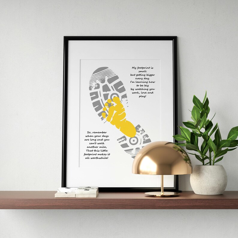 Op de afbeelding: Ingelijste kunstafdruk met een grijs en geel voetstappenontwerp en motiverende tekst. Het kunstwerk bevat een schoenafdruk en een gestileerde hand, met inspirerende zinnen in een modern lettertype. De print wordt tentoongesteld op een plank met een gouden lamp en een plant in pot.