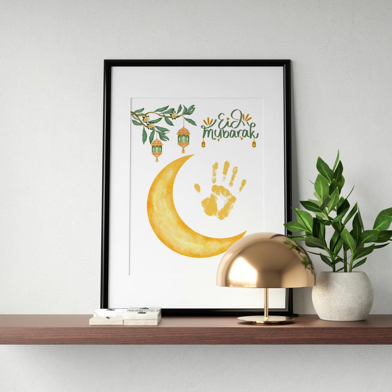 May include: Framed art print featuring a watercolor crescent moon and handprint. The print includes the text "Eid Mubarak" and decorative elements of lanterns and foliage. The artwork is displayed on a shelf with a gold lamp and a potted plant.