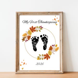 May include: A framed print featuring the text My First Thanksgiving and 2026 in black script. The centre displays two black baby footprints enclosed within a circular gold line, decorated with colourful autumn leaves in orange, yellow, and brown.