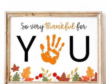 May include: A framed print with the words "So very thankful for YOU" in black and orange. The "Y" and "U" are large black letters, with an orange handprint between them. The bottom of the print has a border of fall leaves and berries.