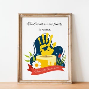 May include: A framed print featuring the text The Saints are our family in heaven. and Happy All Saints Day. The illustration shows a yellow handprint above a yellow candle, surrounded by green leaves, a white flower, and a red ribbon.