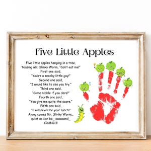 Puede incluir: Una impresión enmarcada con el título "Five Little Apples" y un poema sobre manzanas y un gusano. La obra de arte incluye personajes de manzanas verdes, una huella de mano roja y un gusano de dibujos animados. El marco es de color marrón claro.
