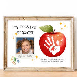 Peut inclure: Impression encadrée avec le texte "My First Day of School". Comprend une photo d'un enfant souriant, une pomme rouge avec une empreinte de main blanche et la citation "On this first day, I leave my mark, A tiny print to light the spark."