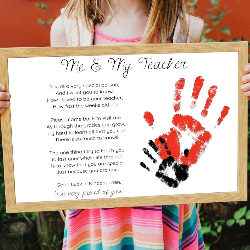 May include: A framed print with the text "Me & My Teacher" and a poem. The print features red and black handprints. The frame is a light brown color. The print is held by a person.