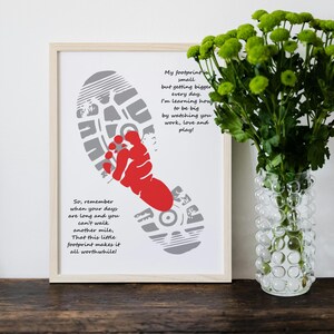 Op de afbeelding: Een ingelijste print met een grijs voetstappenontwerp en een rode babyvoet. De print bevat inspirerende tekst over groei en liefde. Een vaas met groene bloemen staat rechts. De lijst is van licht hout.